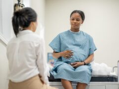 A young pregnant woman sit up on an exam table in her doctors office during a routine prenatal check-up. She wearing a medical gown and her doctor is seated across from her as the two talk.